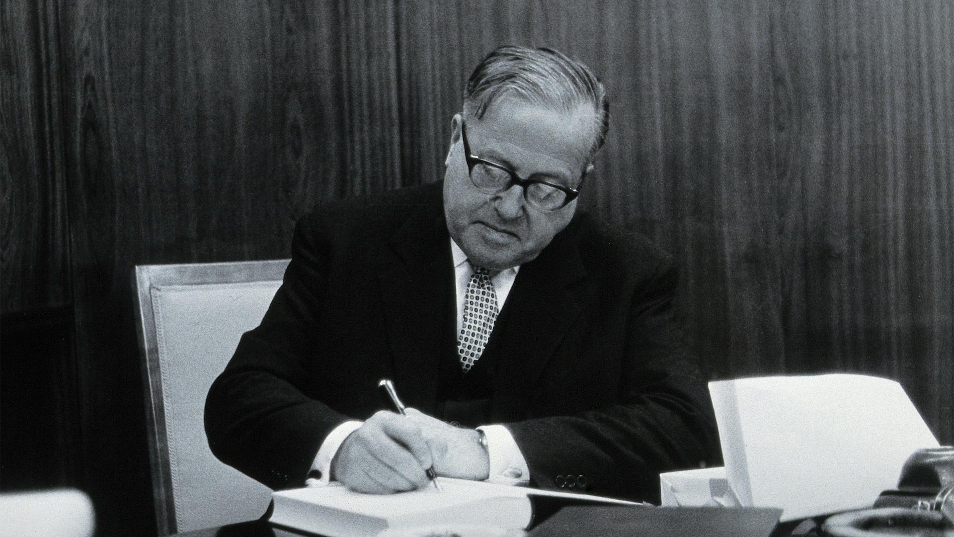 Schwarz-weiss-Aufnahme von Rudolf Geigy am Scheibtisch, während er ein Buch signiert.