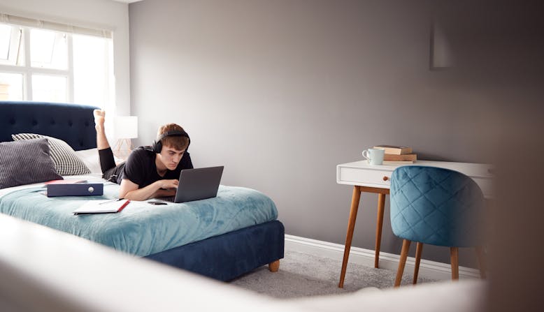 Male college student wearing headphones lies on his bed working on his laptop.
