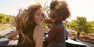 Two women standing up in an open car, looking and smiling at the camera behind them.
