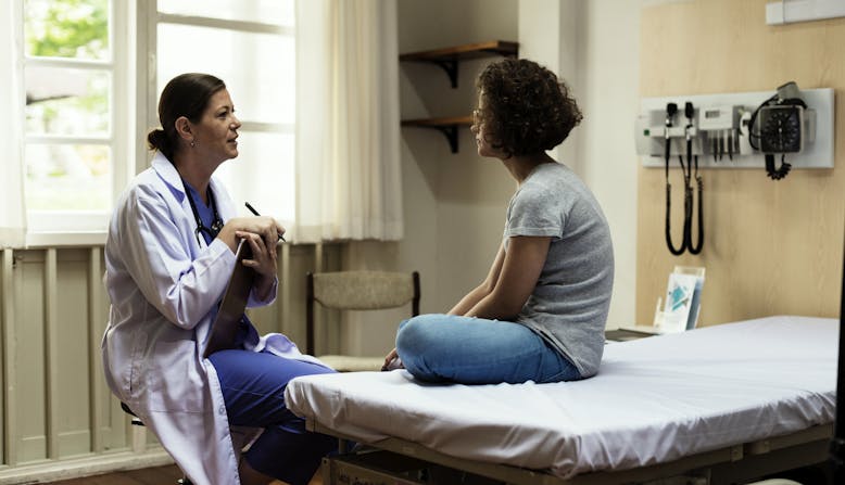 A woman doctor with a clipboard talking to a woman patient sitting on a hospital bed.