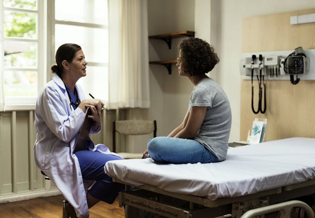 A woman doctor with a clipboard talking to a woman patient sitting on a hospital bed.