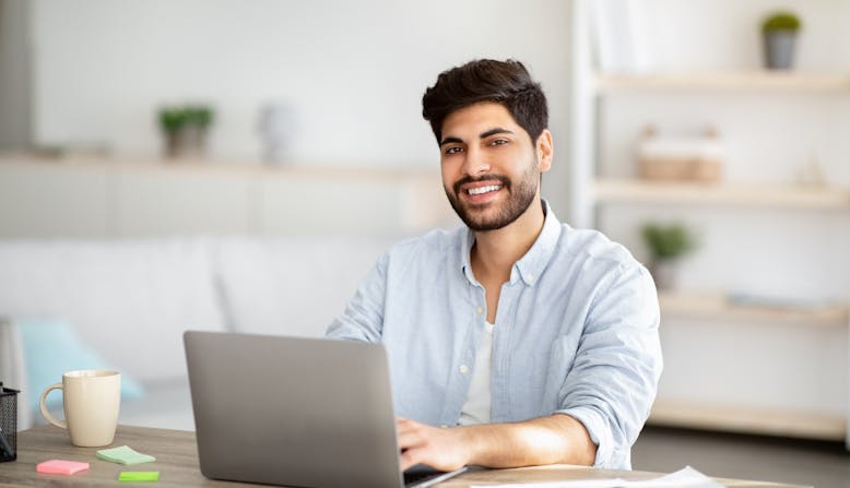 Portrait of happy arab man sitting at desk with laptop, working at home office.