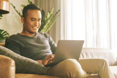 A young asian man sits on a couch looking at a laptop computer.
