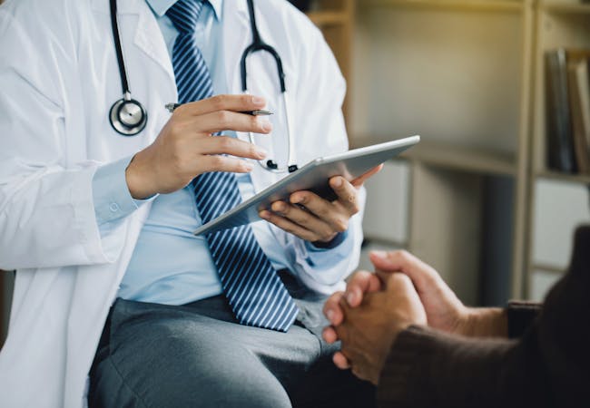 A doctor with a tablet shown from the neck down seated and talking to a patient with clasped hands.