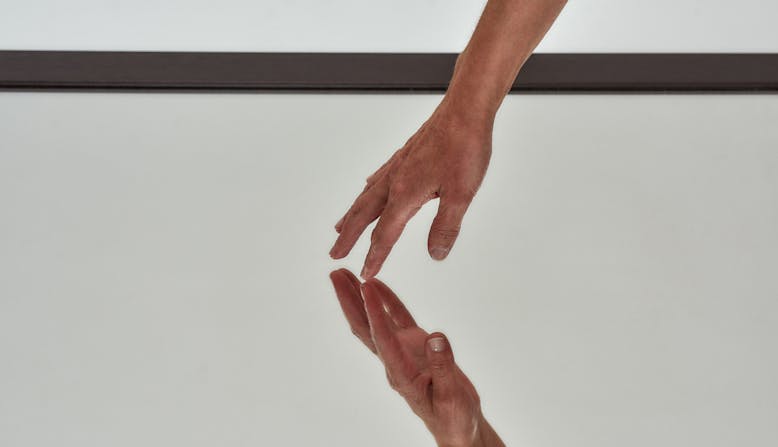 A hand shown touching a mirror with the reflection of the hand seen.