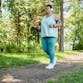 A Black woman runs through a path in a park with trees behind her and headphone on.