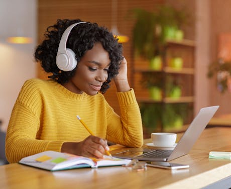 A smiling woman listens to a podcast or webinar while taking notes in a café setting.