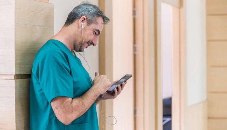 A happy doctor wearing green scrubs using his mobile phone to listen to media in a hospital.