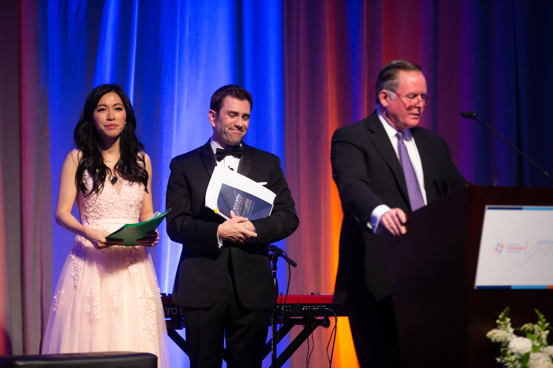 2019 Commit to Cure Gala MCs stand behind Chip Newton as he speaks at the podium.