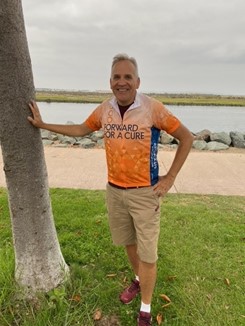 Mark Parikka standing outside with his hand on a nearby tree, smiling, with a "Forward for a Cure" racing shirt on.