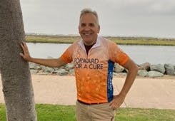 Mark Parikka standing outside with his hand on a nearby tree, smiling, with a "Forward for a Cure" racing shirt on.