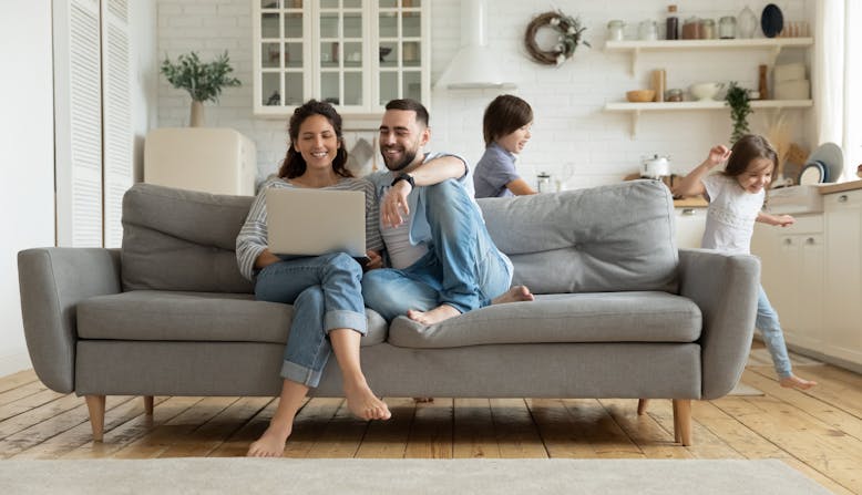 Two parents sit on the couch smiling at a laptop with their two kids playing in the background.