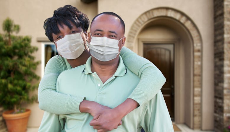 A couple stand together in front of their house smiling and wearing masks.