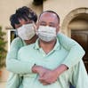 A couple stand together in front of their house smiling and wearing masks.