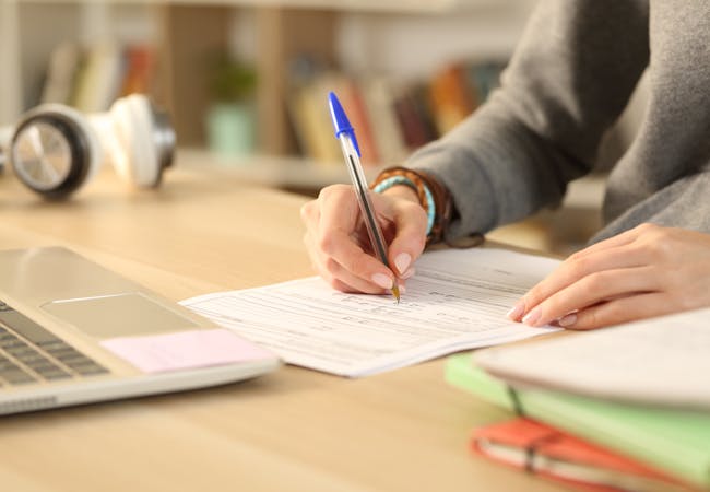 A woman is filling out a paper form with a pen, and books and a computer are in the foreground.