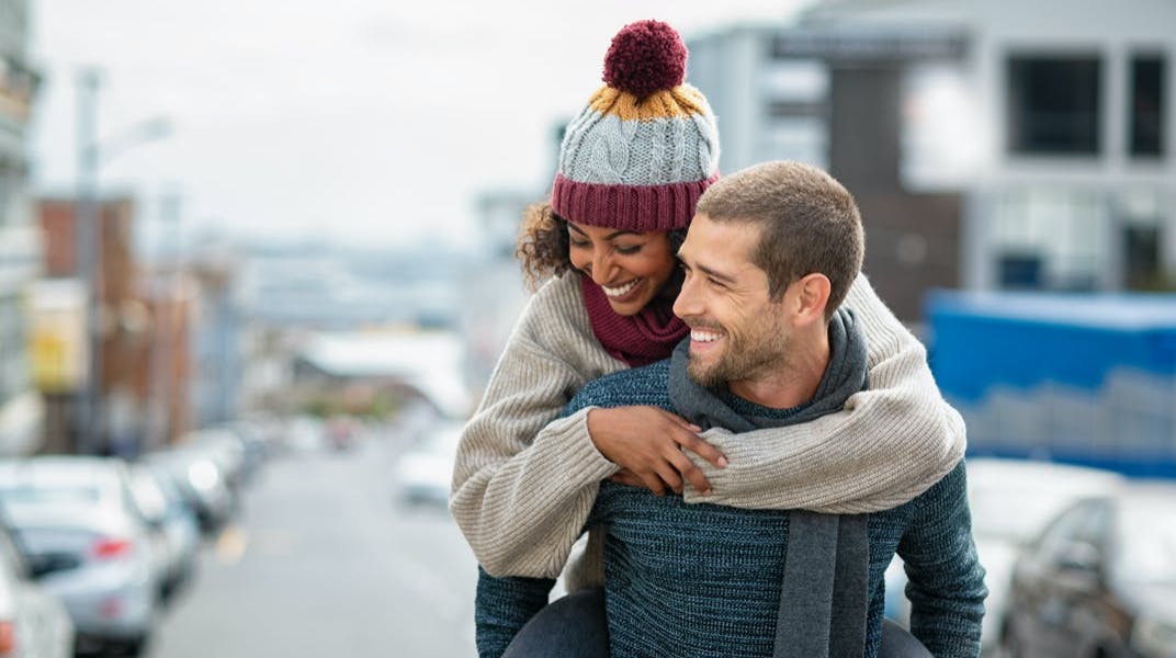 A man gives a lady a piggyback ride, while both are wearing winter clothing. 