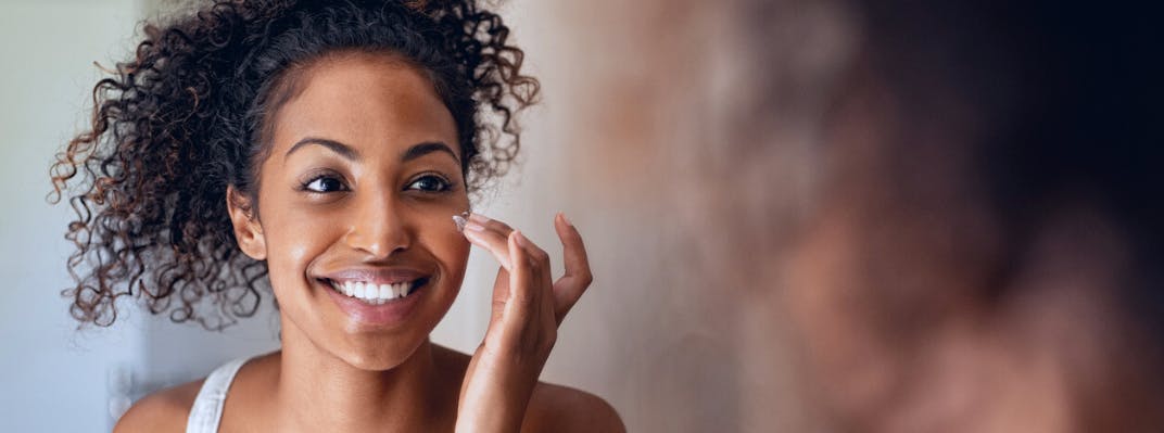 A woman smiles while putting lotion on her face.