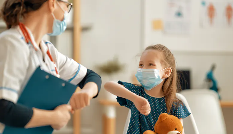 A doctor and child patient bump elbows and smile in a clinic.