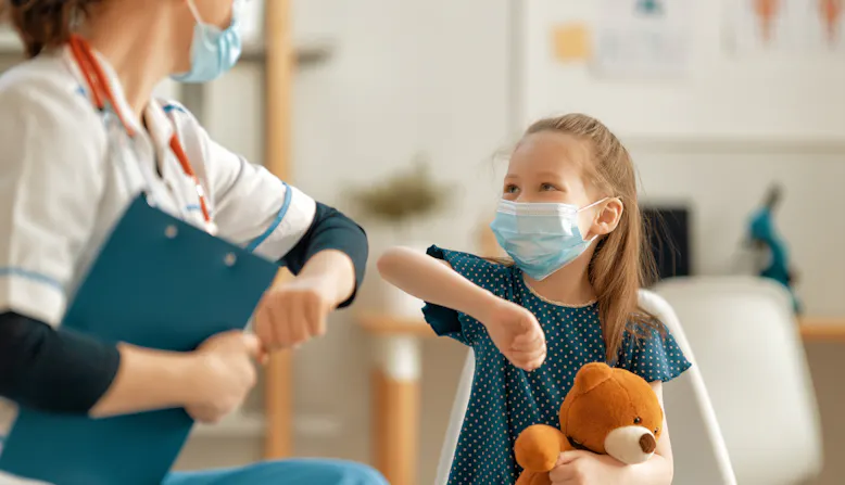 A doctor and child patient bump elbows and smile in a clinic.