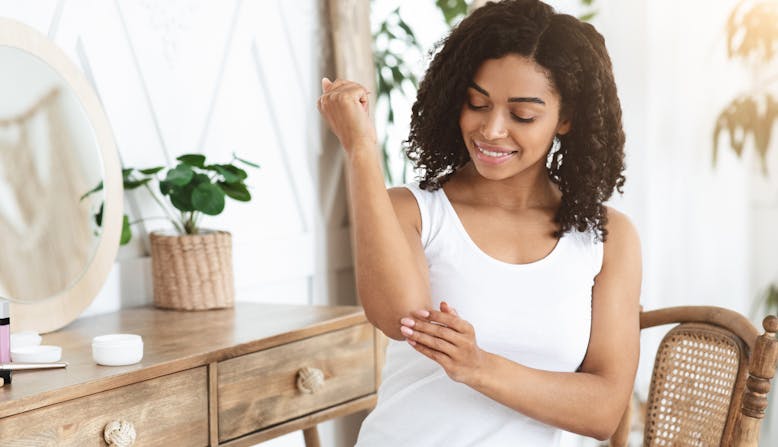 A woman sits next to a vanity and puts cream on her skin.
