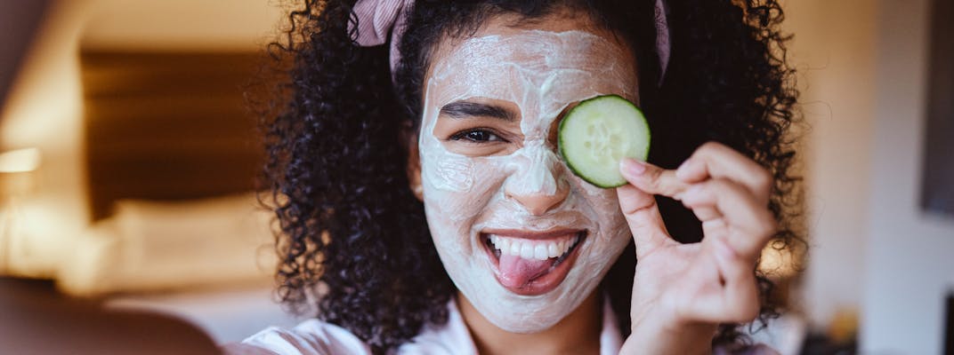 A girl with dark curly hair takes a selfie with a skincare face mask on holding a cucumber and sticking her tongue out.