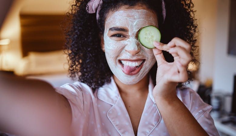A girl with dark curly hair takes a selfie with a skincare face mask on holding a cucumber and sticking her tongue out.