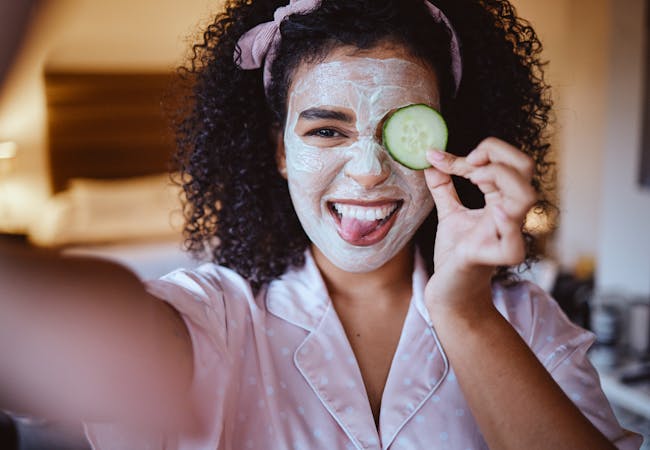 A girl with dark curly hair takes a selfie with a skincare face mask on holding a cucumber and sticking her tongue out.