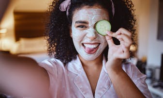 A girl with dark curly hair takes a selfie with a skincare face mask on holding a cucumber and sticking her tongue out.