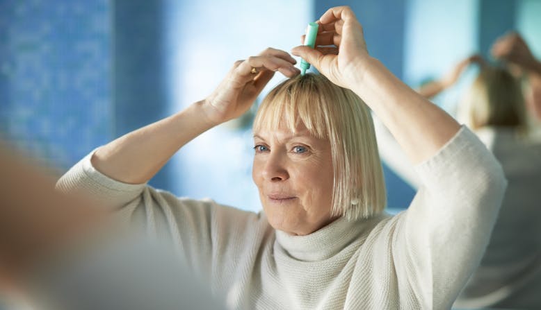 A woman looks in her bathroom mirror while applying treatment to her scalp.
