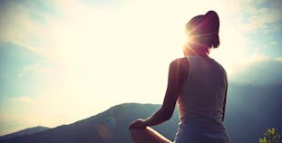 A woman sitting legs crossed with a sunrise and mountains in the background.