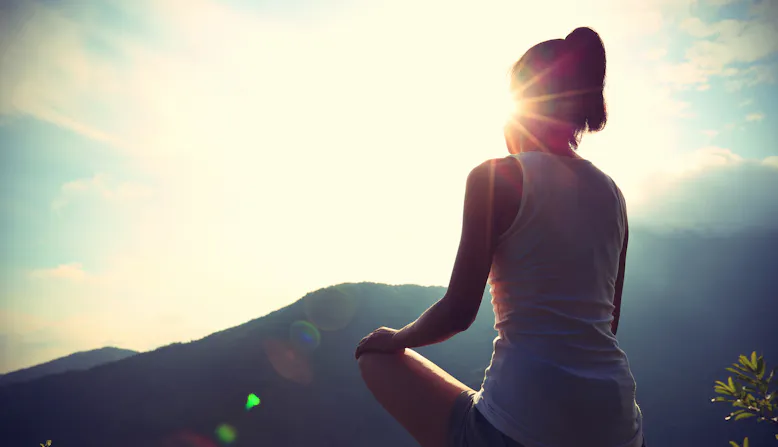 A woman sitting legs crossed with a sunrise and mountains in the background.