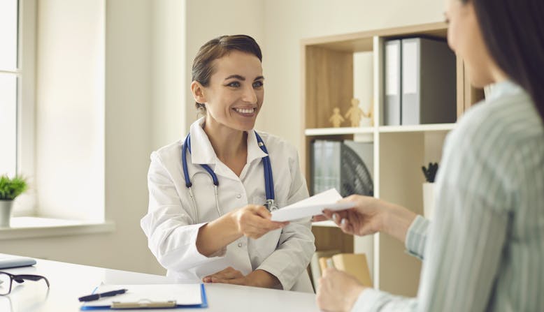 A smiling doctor hands her patient a prescription in an office.