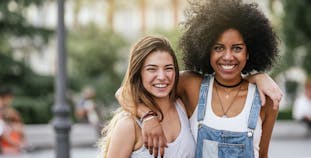 Two women embracing on the sidewalk in a city with trees in the background.