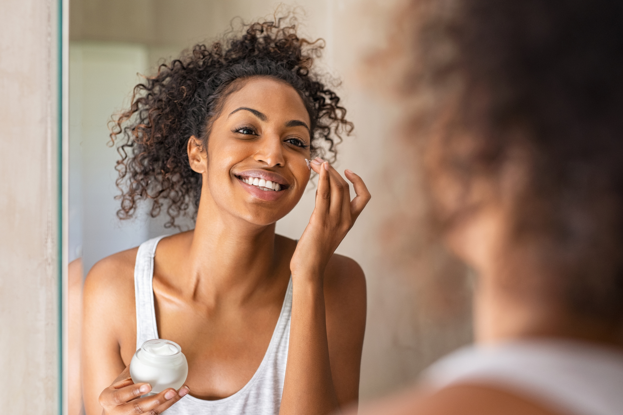 A woman with darker skin looking in the mirror and applying lotion to her face.