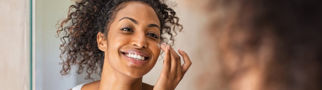 A woman with darker skin looking in the mirror and applying lotion to her face.