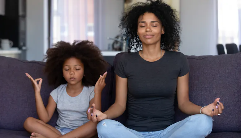 A mother sits on the couch with her daughter meditating.