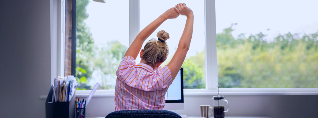 A young woman sits at her desk and stretches with her arms over her head.