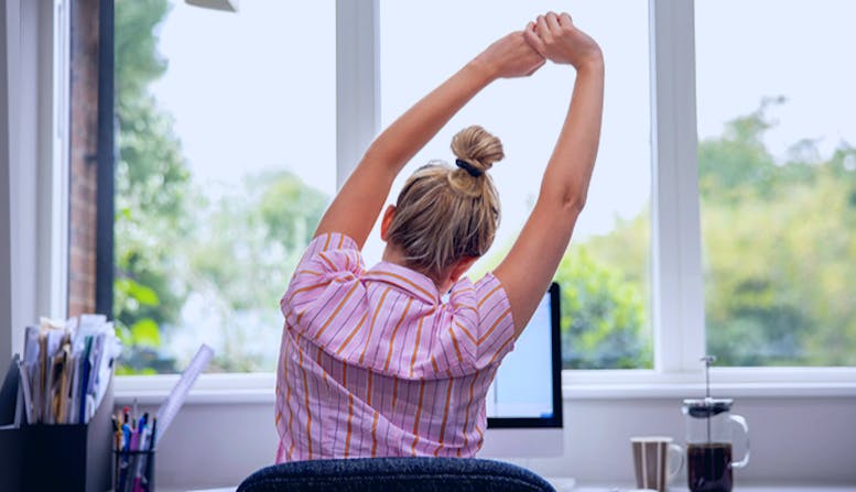 A young woman sits at her desk and stretches with her arms over her head.