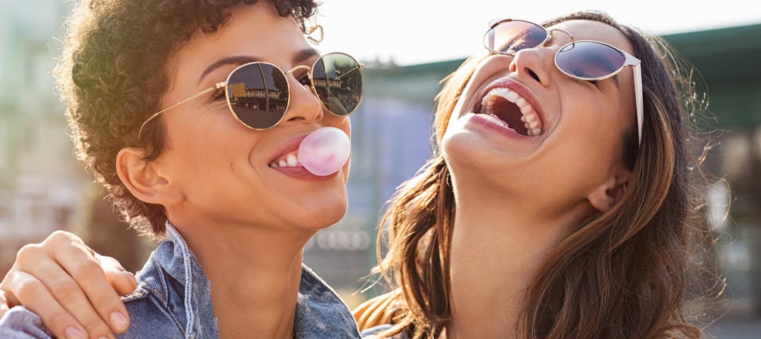 Two women, one blowing a bubble with gum, hug outside.