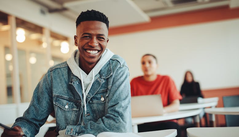 A young black man sitting in a classroom setting smiling with a book.