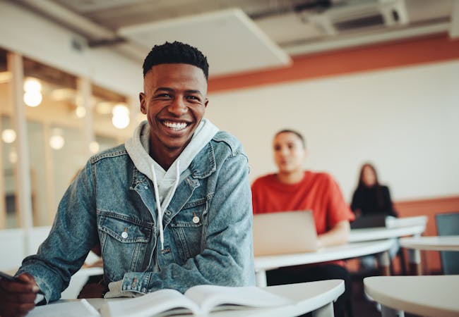 A young black man sitting in a classroom setting smiling with a book.