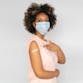 A black woman with an afro wearing a mask and pointing at a band-aid on her upper arm.