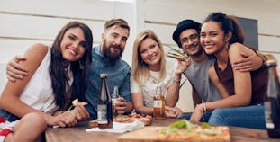 A multiracial group of friends sitting at a dinner table with food and drinks, smiling at the camera.