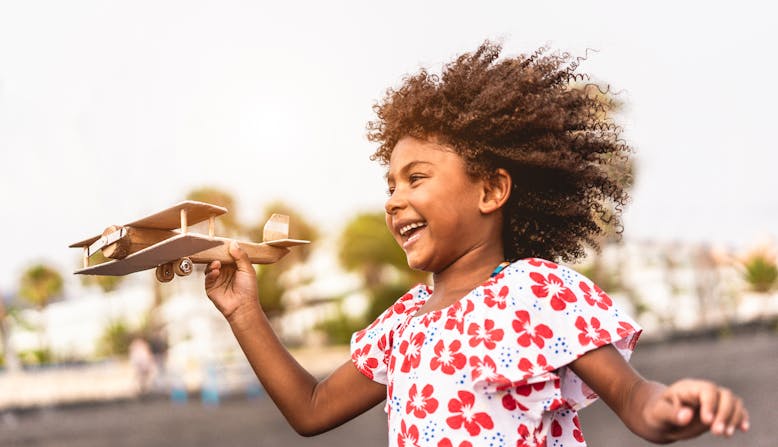 A black girl running outside with a wooden toy plane in her hand.
