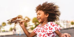 A black girl running outside with a wooden toy plane in her hand.
