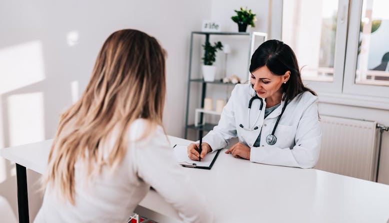 A female doctor writes notes while interacting with a female patient in an office.