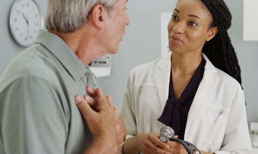 A female doctor listens as a patient speaks and holds his chest.