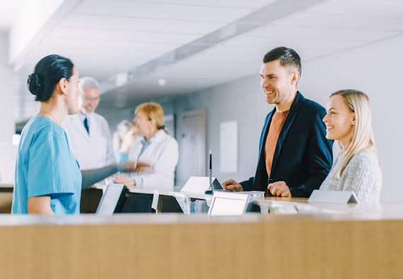 A couple stands at a receptionist desk and talks to a health care professional.  