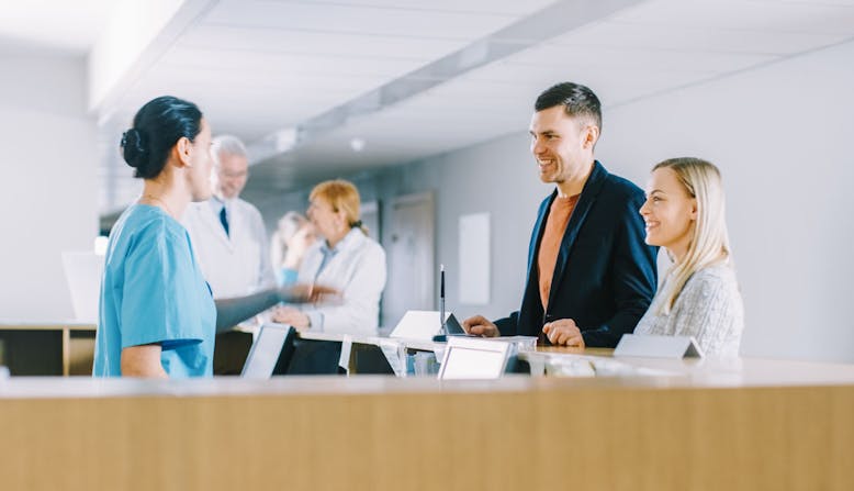 A couple stands at a receptionist desk and talks to a health care professional.