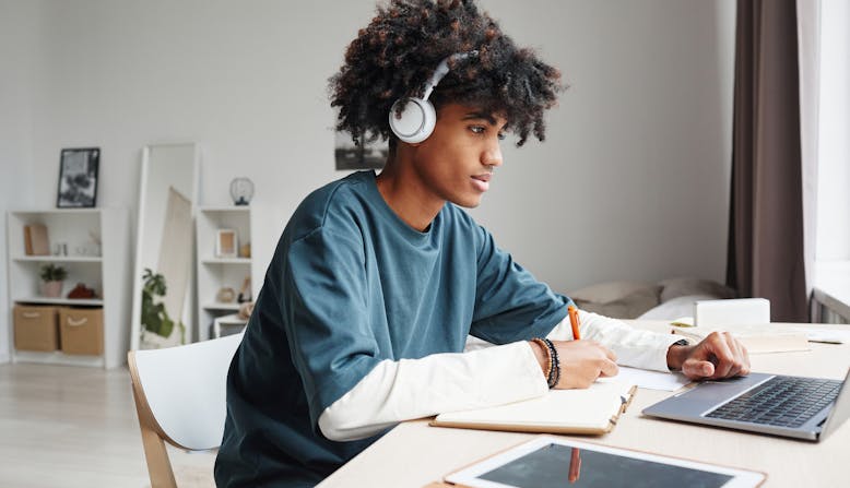 A young man sits at a desk in a bedroom taking notes from his laptop computer.